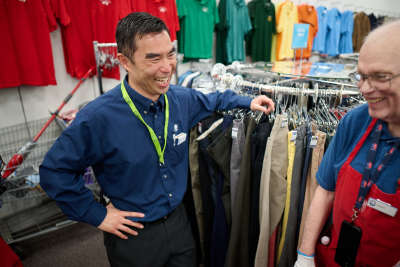 thrift store supervisor smiles as he talks with a thrift store worker who is putting clothes on the retail floor